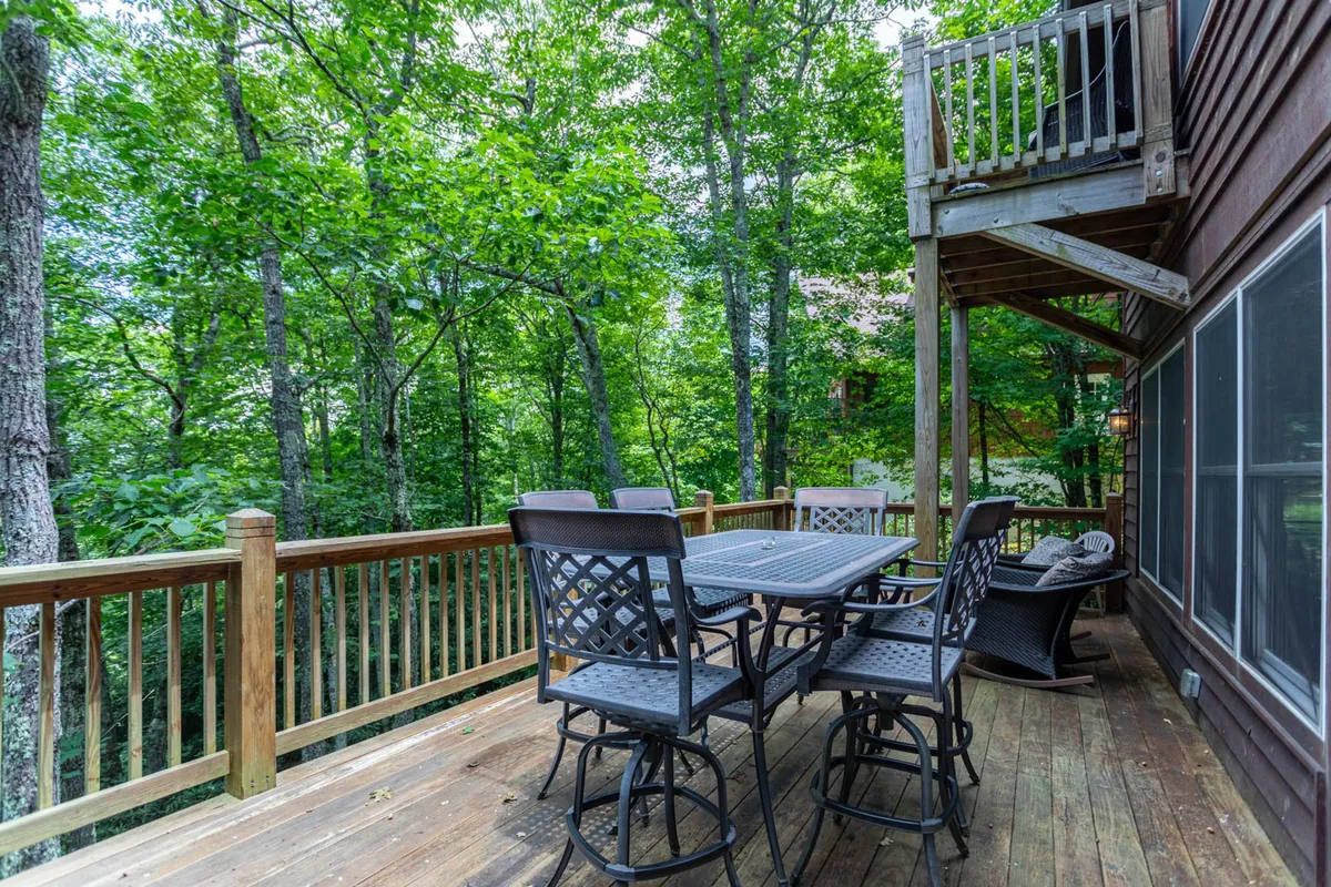 An inviting deck with an outdoor dining set, surrounded by lush greenery and mountain views visible past the railing. The space is perfect for outdoor meals, enjoying fresh air, and relaxing with nature all around.